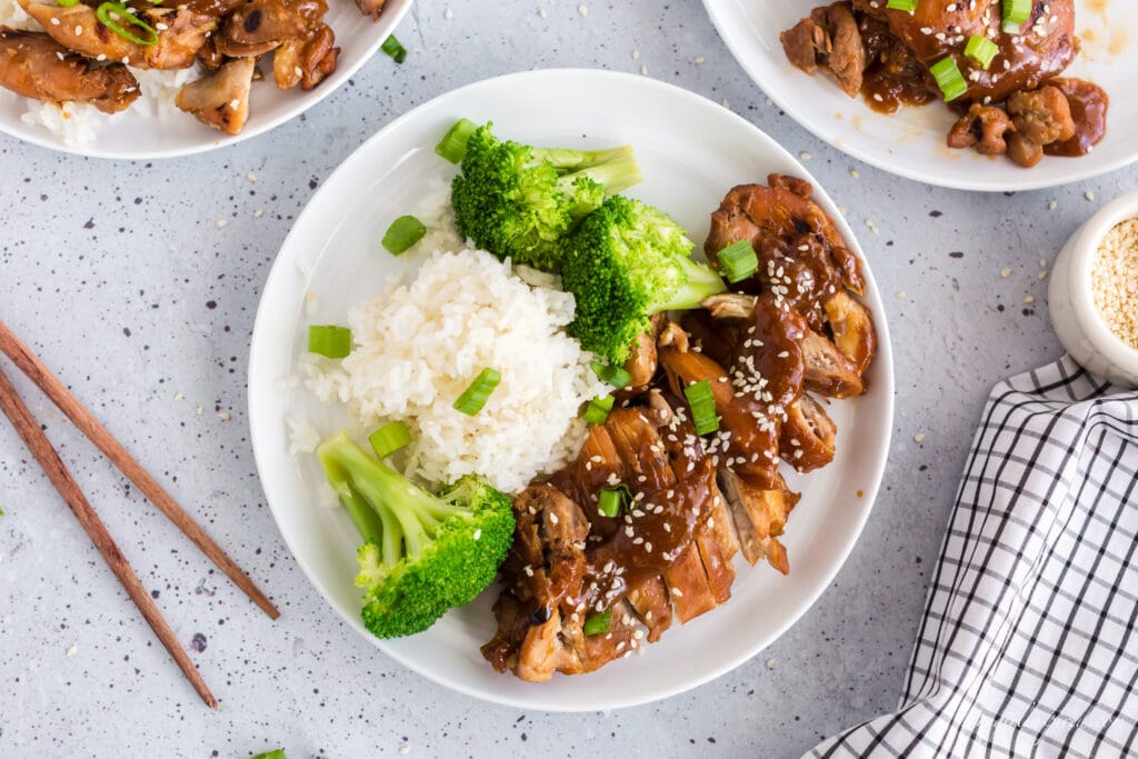 overhead view of teriyaki chicken on white plate with broccoli and rice, sesame seeds and green onion garnish