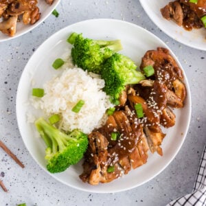 overhead view of teriyaki chicken on white plate with broccoli and rice, sesame seeds and green onion garnish