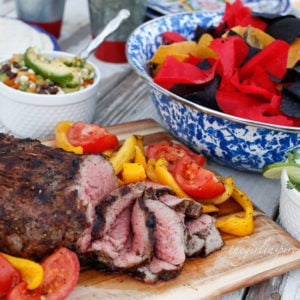 Grilled and sliced Chipotle steak on wooden cutting board with veggies, paper plates and utensils, bowl of red and blue tortilla chips