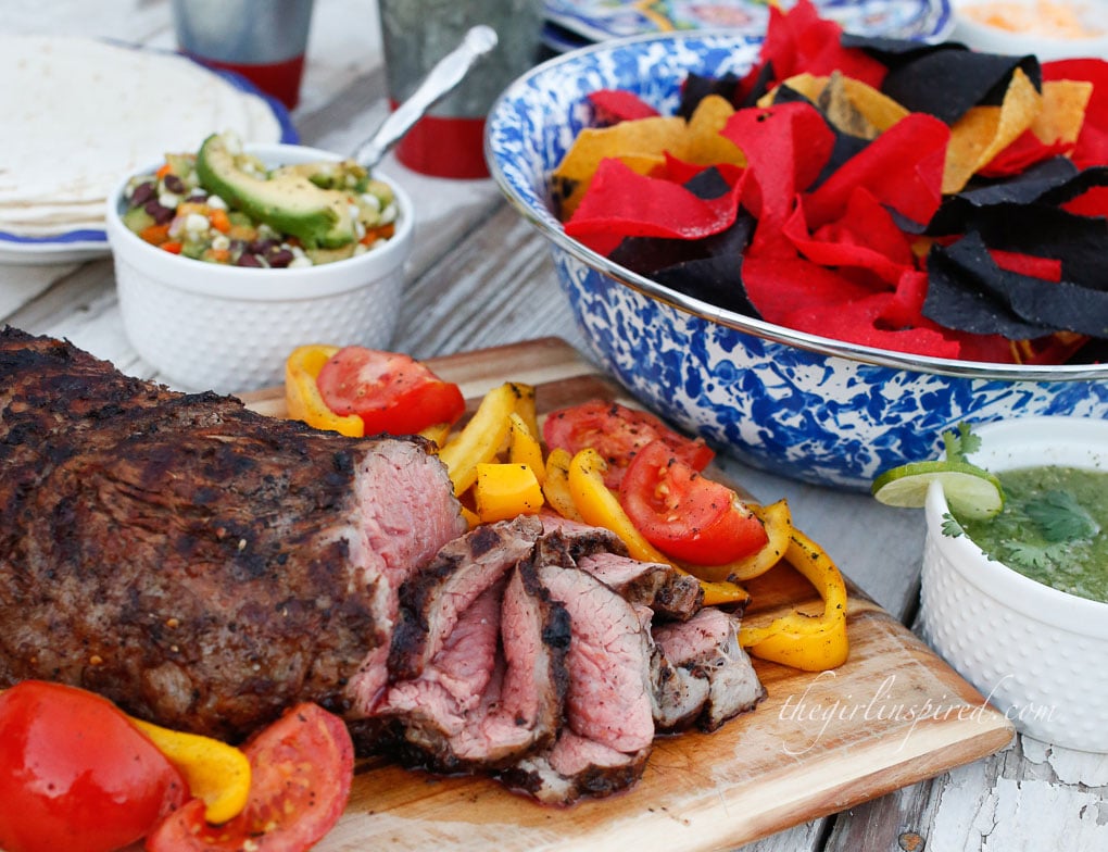 grilled and sliced tri tip on wooden cutting board with veggies, paper plates and utensils, bowl of red and blue tortilla chips