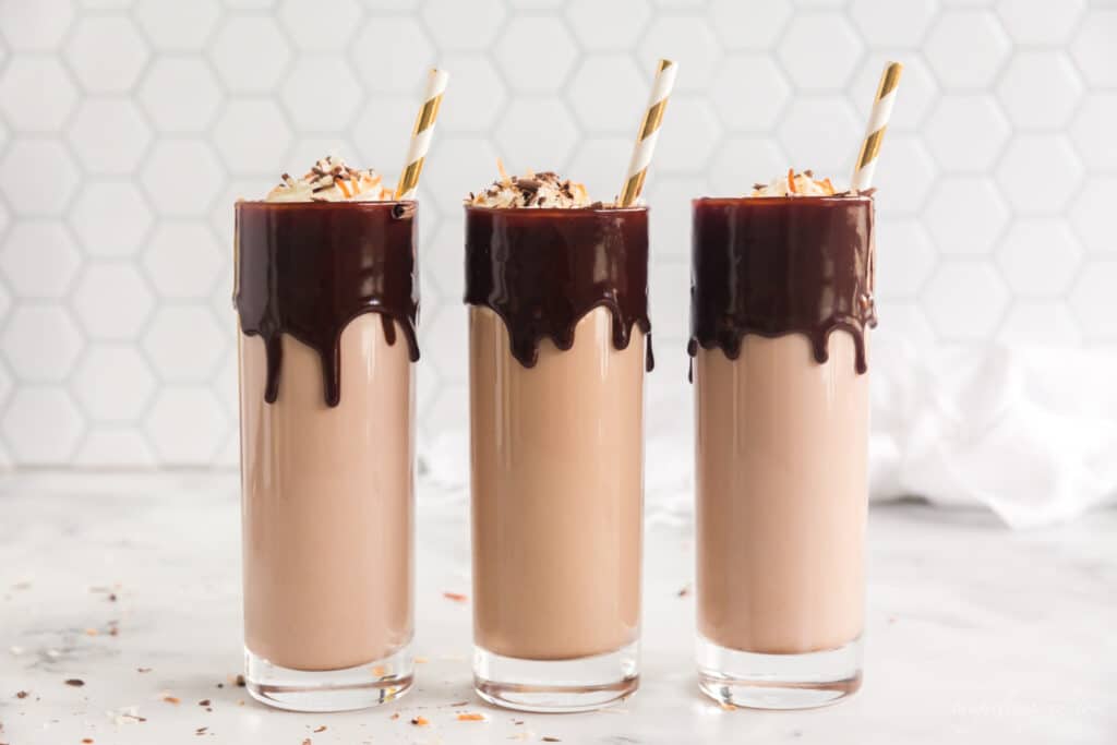 three tall glasses with chocolate dripping from rim, filled with chocolate milk and liquor and topped with whipped cream and gold stripes straws