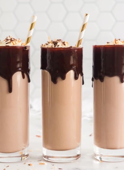 three tall glasses with chocolate dripping from rim, filled with chocolate milk and liquor and topped with whipped cream and gold stripes straws