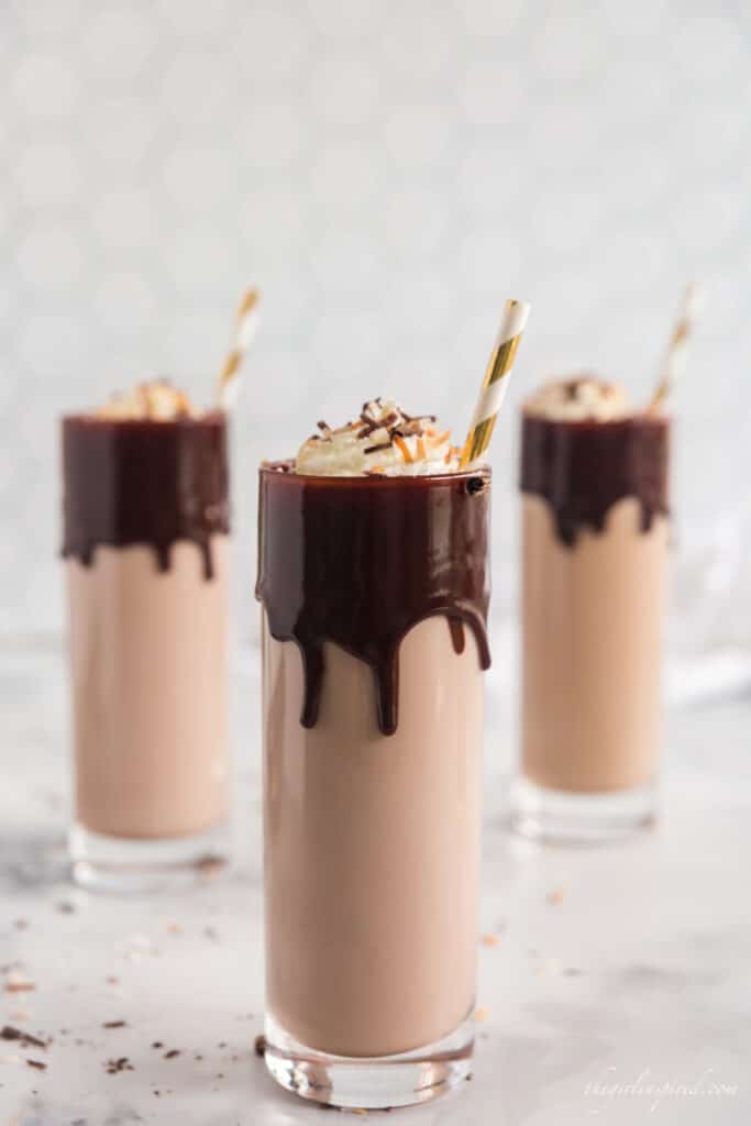three tall glasses of coconut rum cocktail with chocolate dripping from rim, filled with chocolate milk and liquor and topped with whipped cream and gold stripes straws