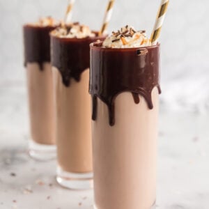 three tall glasses with chocolate dripping from rim, filled with chocolate milk and liquor and topped with whipped cream and gold stripes straws