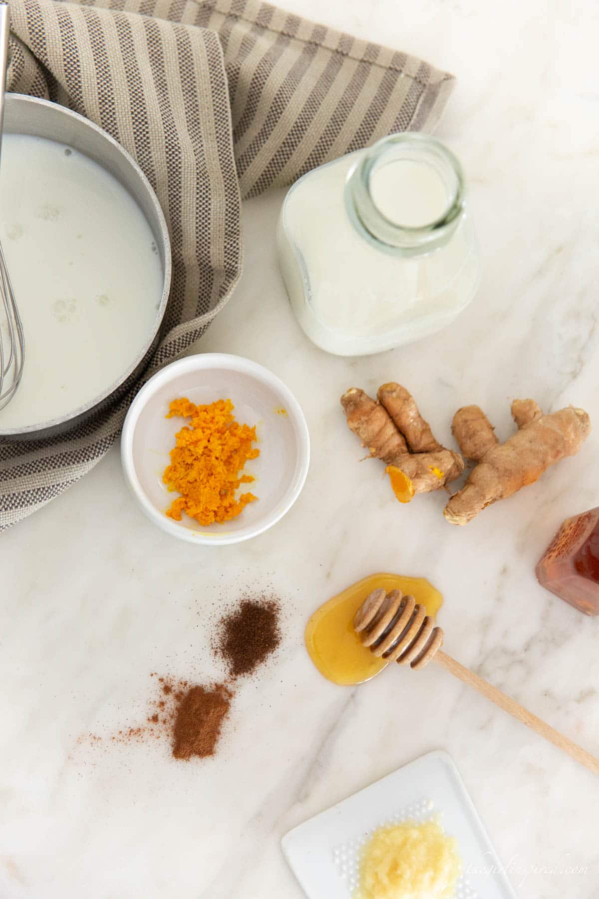 ingredients including grated and whole turmeric, honey, cinnamon and clove spices, a glass jar of milk, dish towel and saucepan with milk and a whisk