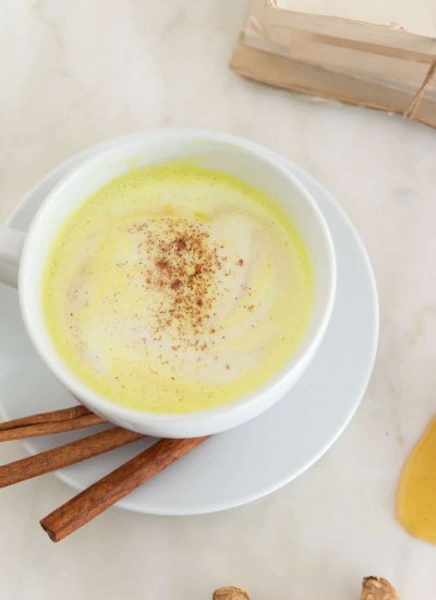 frothy yellow milk in white mug with cinnamon sticks, whole turmeric root, cinnamon sticks, and honey