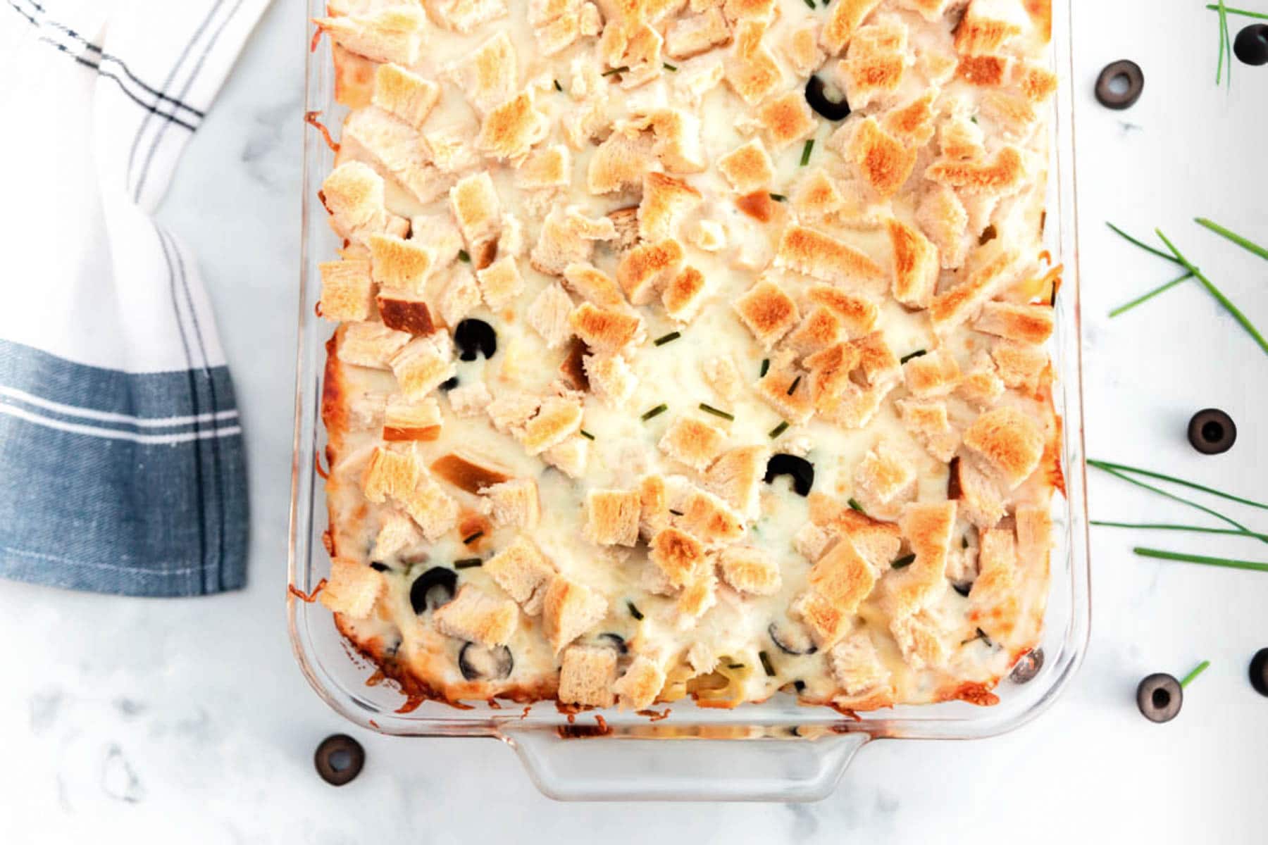 baked tuna casserole in glass baking dish with kitchen towel, chives and sliced olives on countertop