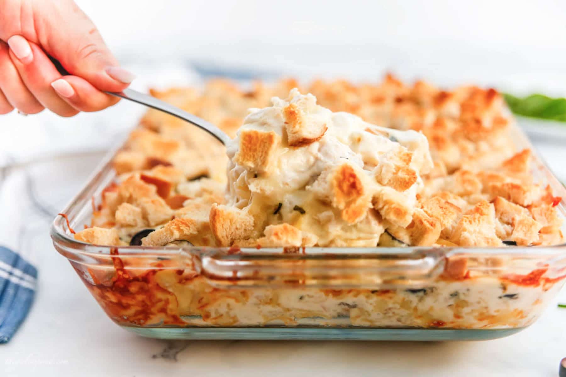 spoonful of baked tuna casserole in glass baking dish with kitchen towel, chives and sliced olives on countertop