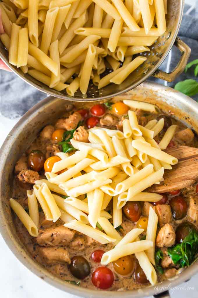 penne pasta pouring into chicken and tomato saucepan mixture