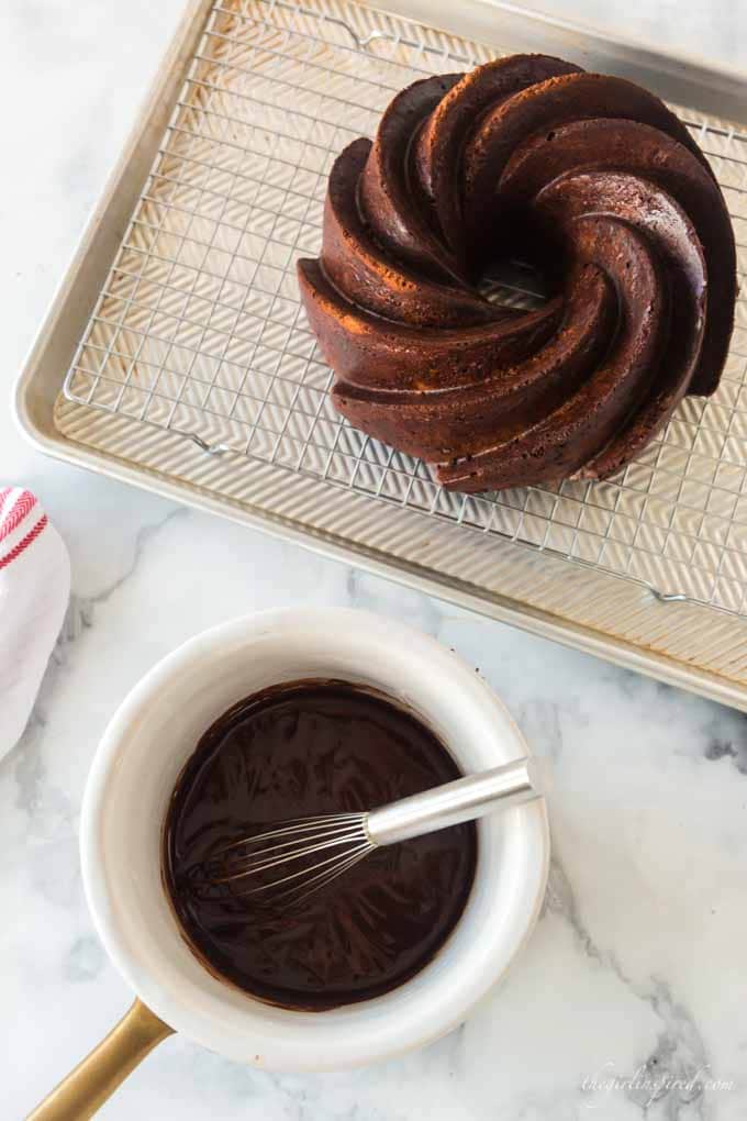 chocolate cake on cooling rack and baking sheet with white pot of melted chocolate glaze and whisk