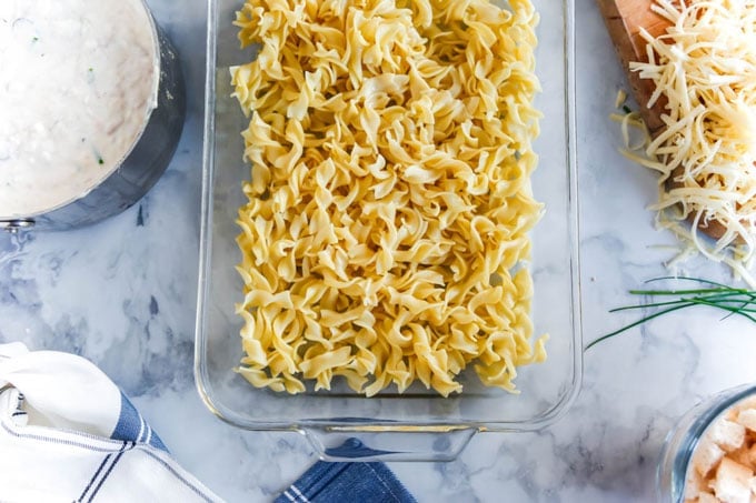 noodles in glass baking dish, grated cheese, chives, white sauce in pan, and dish towel set out beside casserole dish