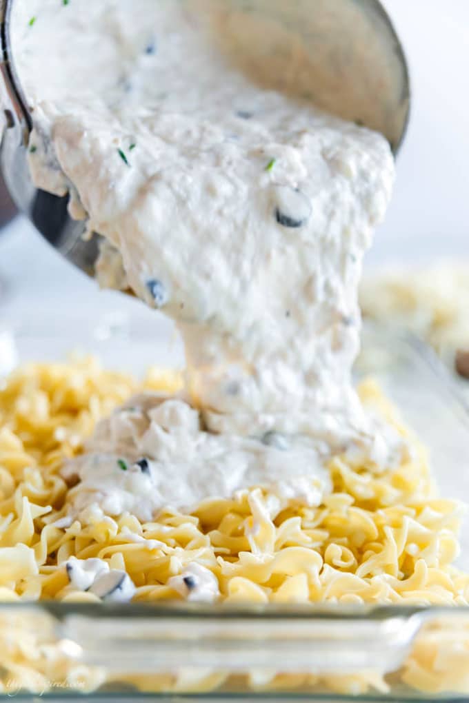 noodles in glass baking dish with white sauce being poured from saucepan over the top