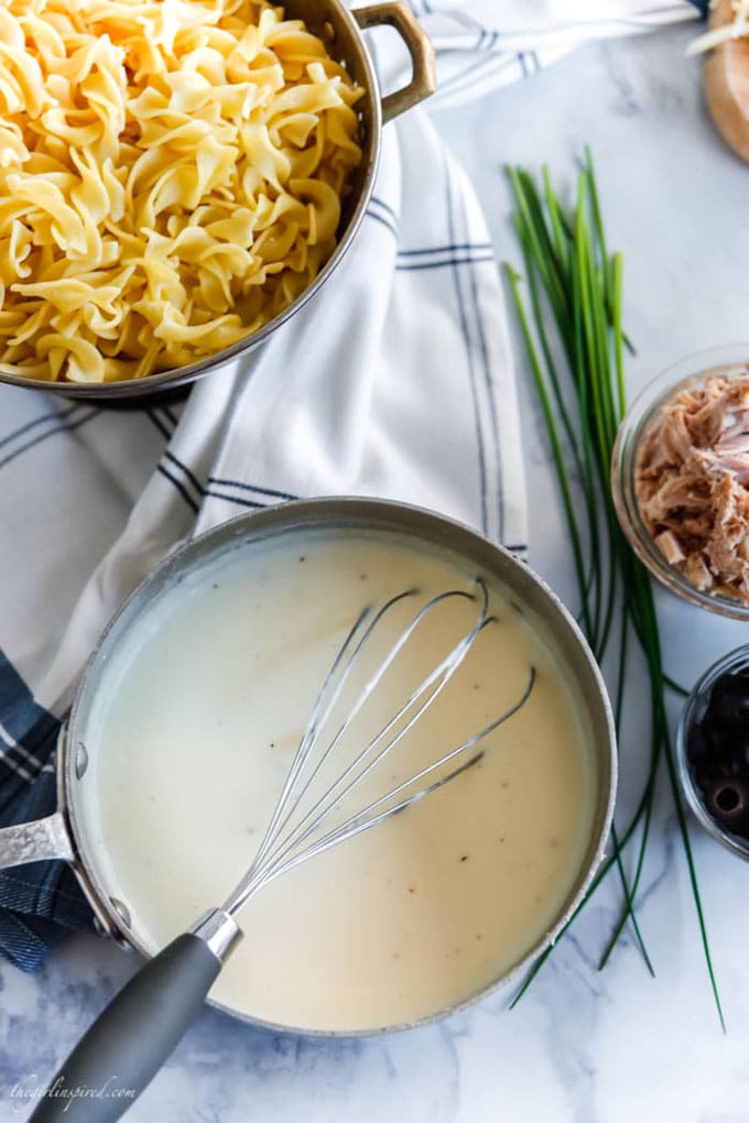 overhead view of saucepan with white sauce, colander with noodles, chives, tuna, and olives in dishes