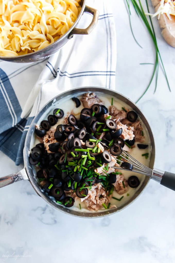 chives, olives and tuna being stirred into white sauce, noodles in colander and dish towel on countertop