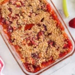 Baked Strawberry Rhubarb Crisp in glass dish with fresh strawberries and rhubarb and red and white linen.