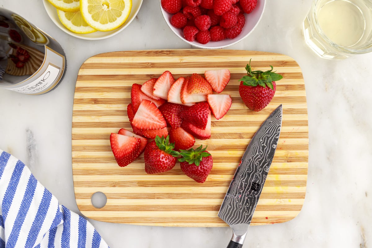 Sliced strawberries on a cutting board, knife, bowl of raspberries and lemon slices.