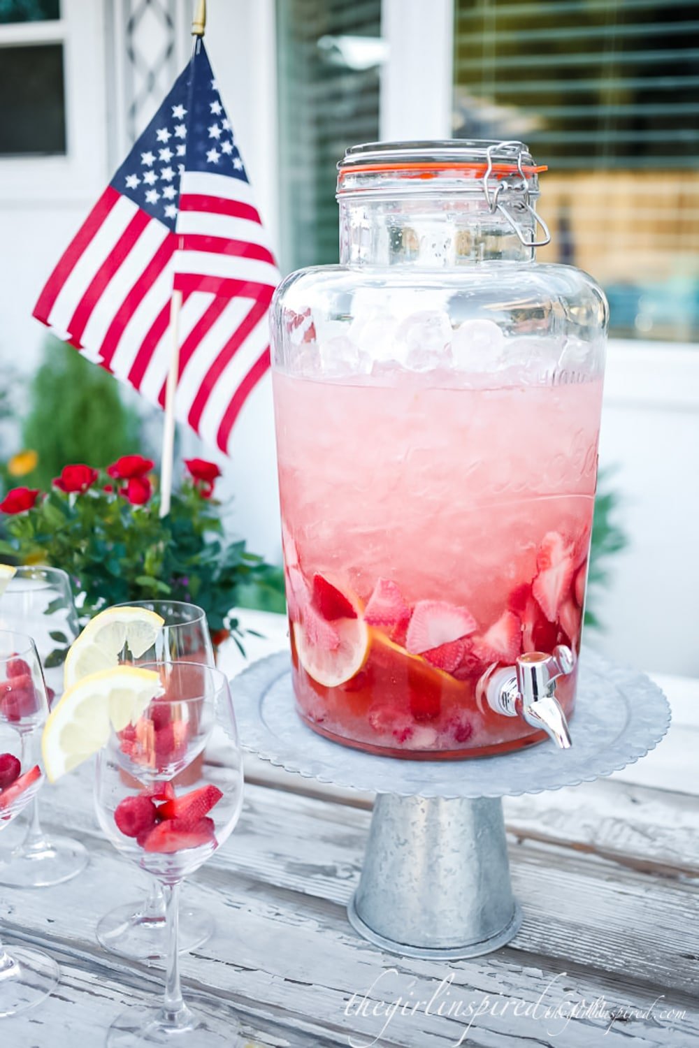 strawberry sangria in large glass drink dispenser on pedestal stand, lemon wedges and fresh berries in wine glasses on table and American flag in the background