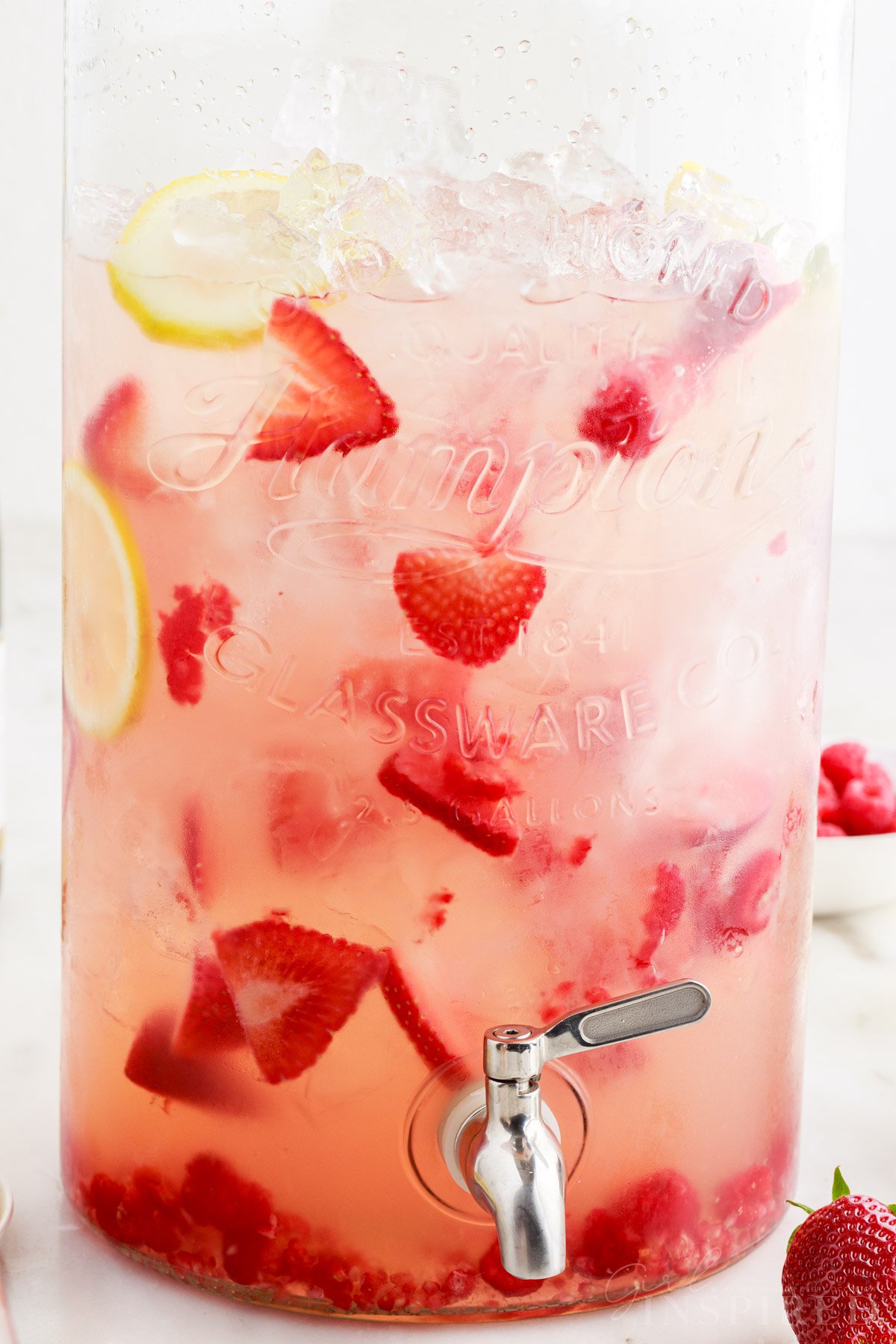 Large glass beverage dispenser full of strawberry sangria with fresh strawberries, raspberries, and lemon slices floating in it.