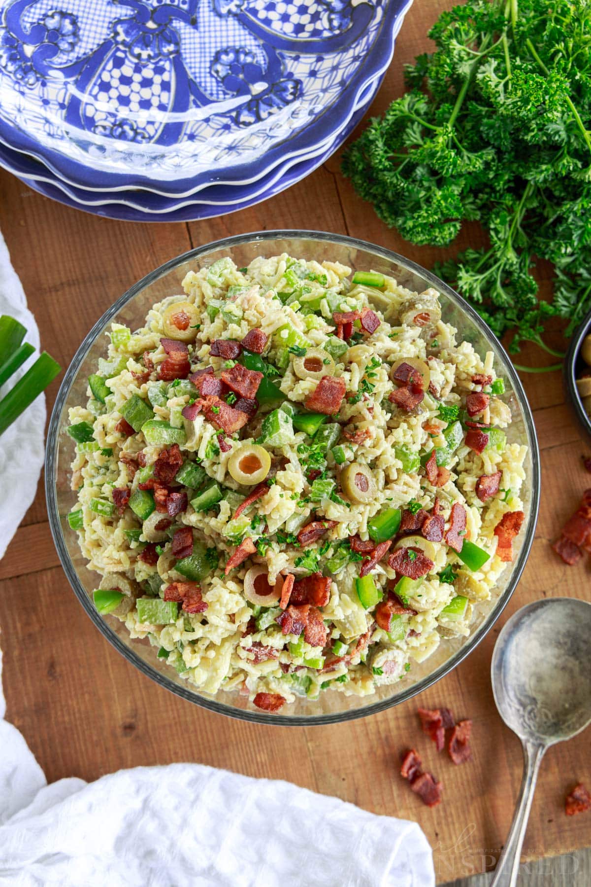 Prepared bowl of Rice a roni salad on a wooden board with stack of plates, bunch of parsley and green onions, and a spoon and linen.