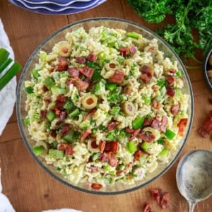Prepared bowl of Rice a roni salad on a wooden board with stack of plates, bunch of parsley and green onions, and a spoon and linen.