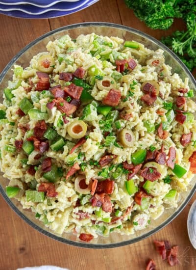 Prepared bowl of Rice a roni salad on a wooden board with stack of plates, bunch of parsley and green onions, and a spoon and linen.