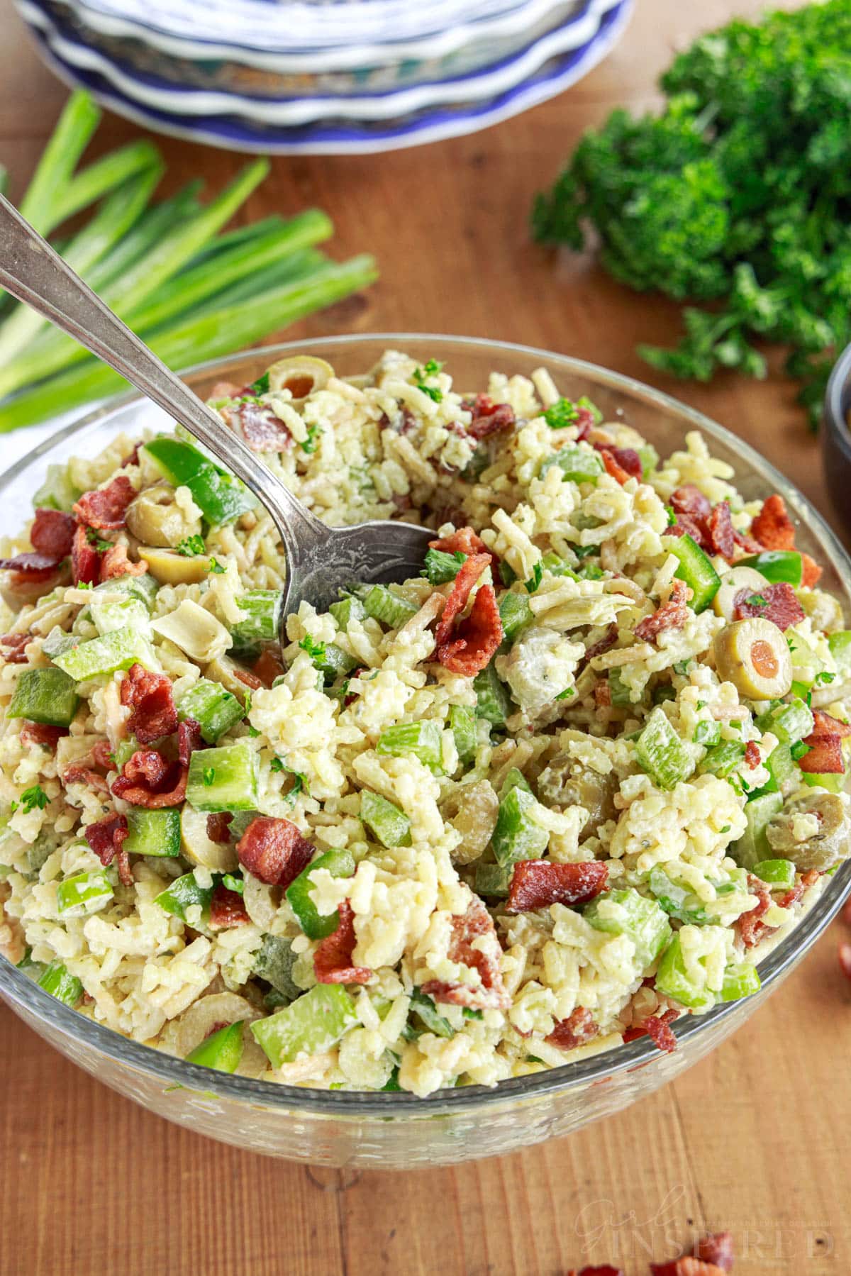 Spoon scooping into prepared bowl of Rice a roni salad on a wooden board with stack of plates, bunch of parsley and green onions.