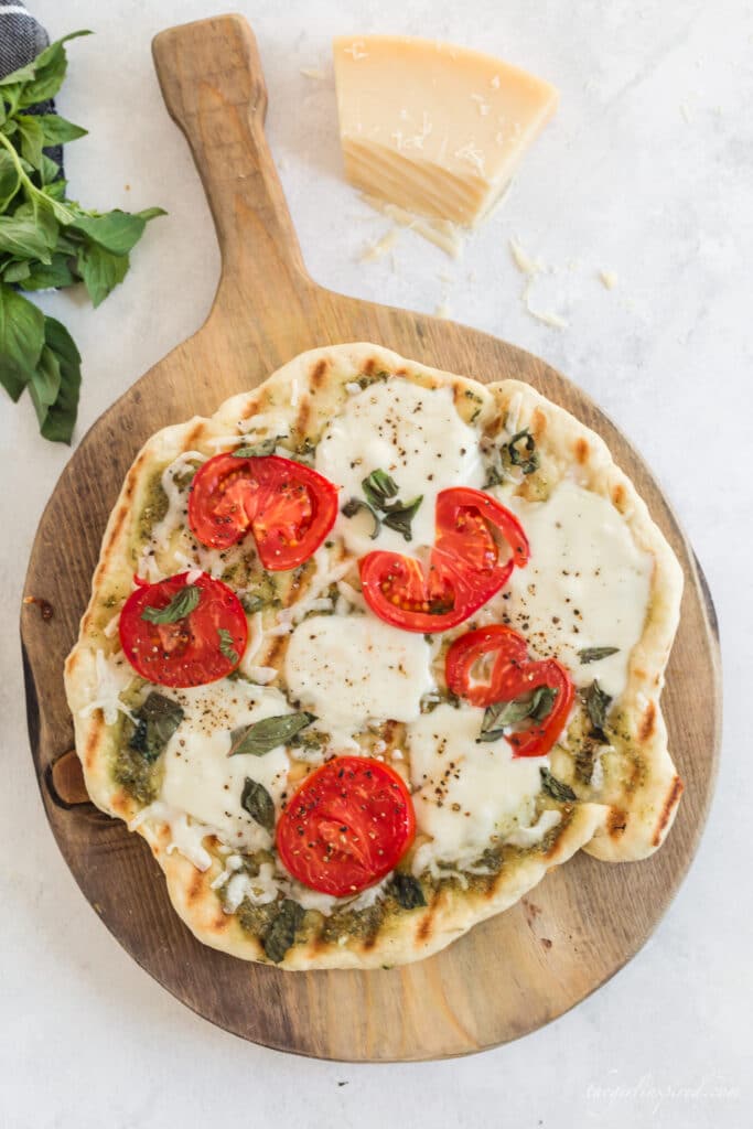 grilled pizza with tomatoes, basil, and melted cheese on wooden board with parmesan, basil leaves, and grey linen in background