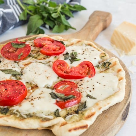 pizza with grill lines, tomatoes, basil, and melted cheese on wooden board with parmesan, basil leaves, and grey linen in background