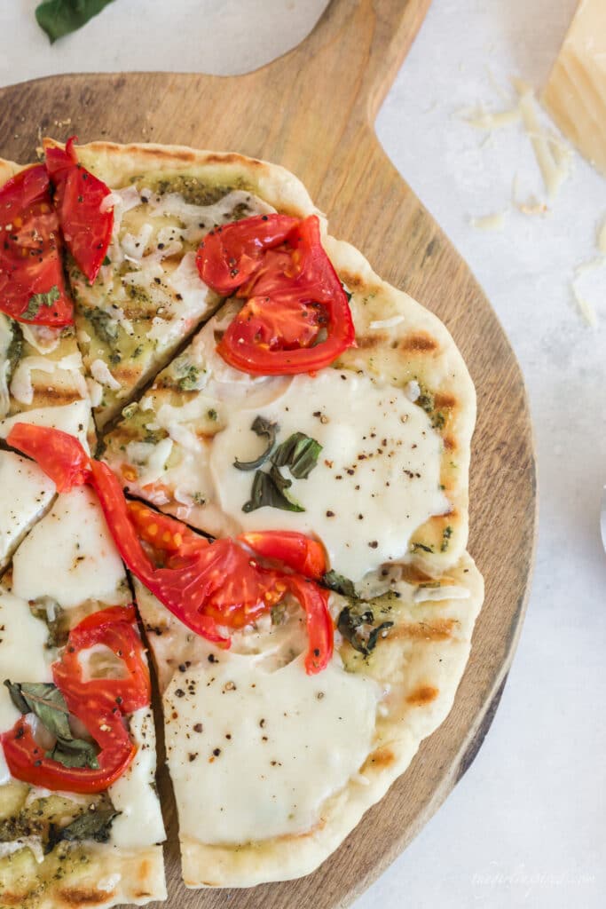 pizza with grill lines, tomatoes, basil, and melted cheese on wooden board with parmesan, basil leaves, and grey linen in background