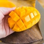 Half of a mango held in a hand with cubes of flesh attached to the skin.