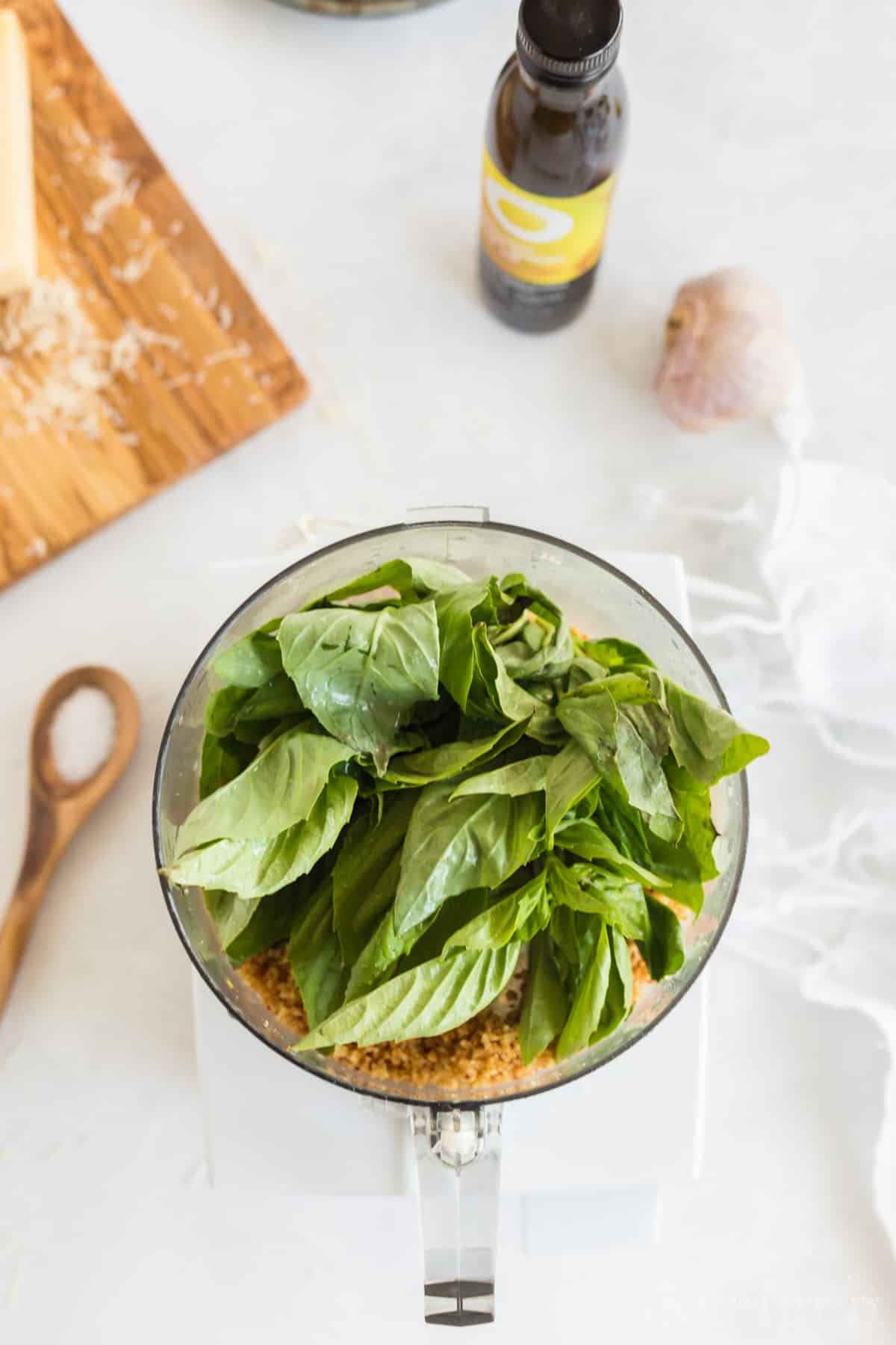 Fresh basil leaves mounded over pine nut mixture in bowl of a food processor.