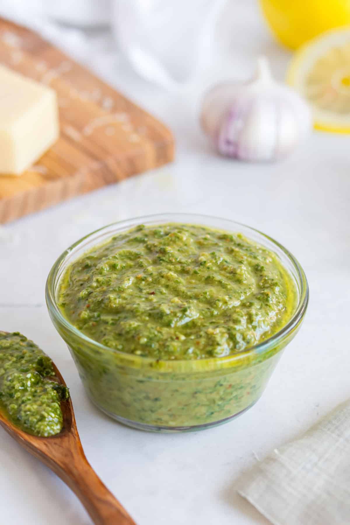 Glass dish filled with lemon pesto, wooden spoon, cutting board with parmesan cheese, lemons, and head of garlic.