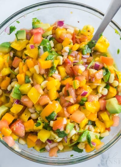 overhead photo of mango avocado salsa in clear glass bowl with whole mangos, lime slices, cilantro, and a red and white linen in background