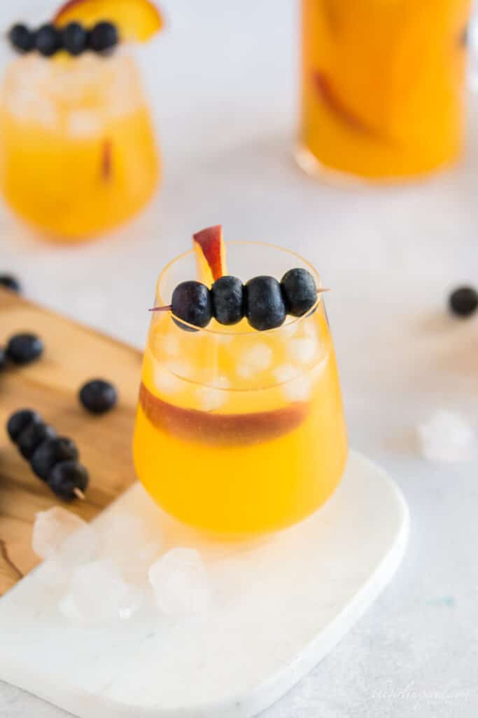 peach sangria in a carafe and wine glasses with blueberries on a toothpick and a peach slice for garnish, marble and wood cutting board
