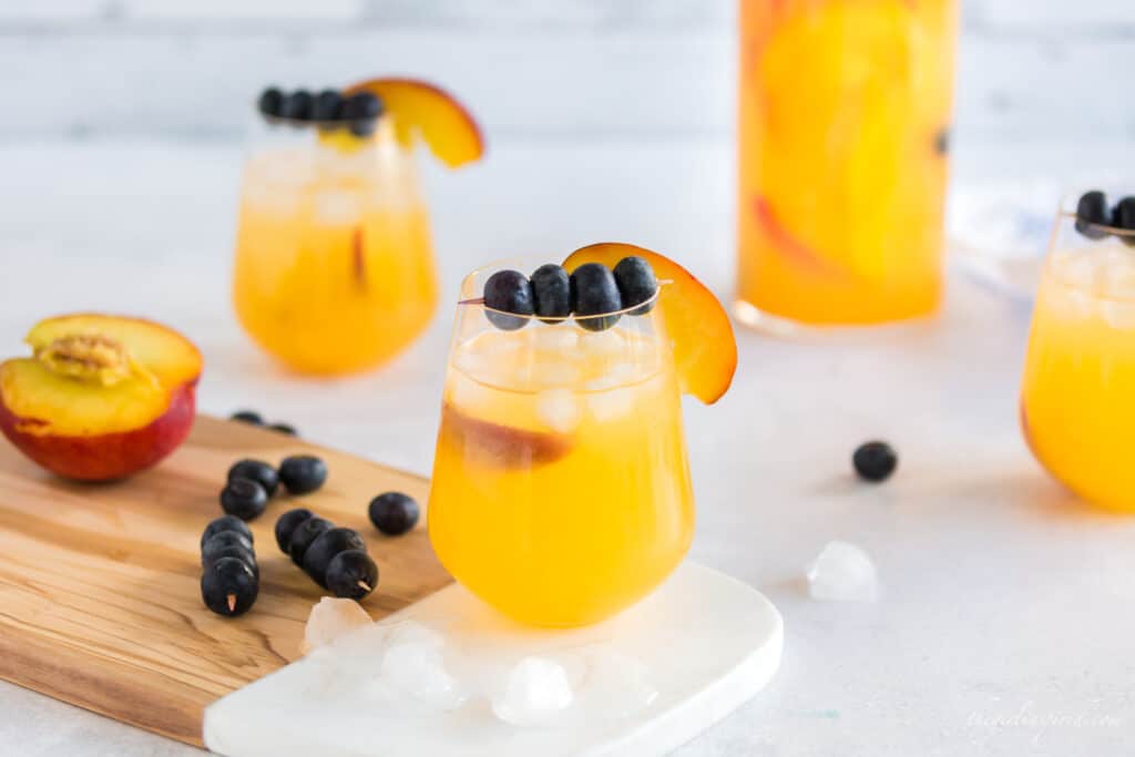 peach sangria in a carafe and wine glasses with blueberries on a toothpick and a peach slice for garnish