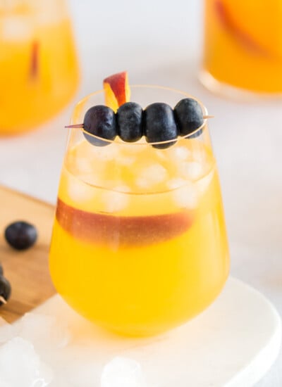 Peach sangria in a carafe and wine glasses with blueberries on a toothpick and a peach slice for garnish.