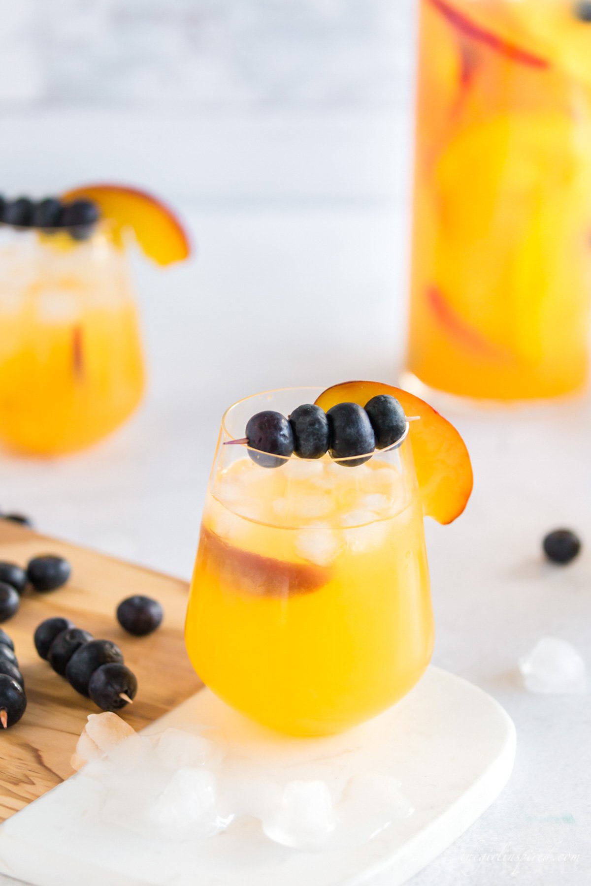 Peach sangria in a carafe and wine glasses with blueberries on a toothpick and a peach slice for garnish.