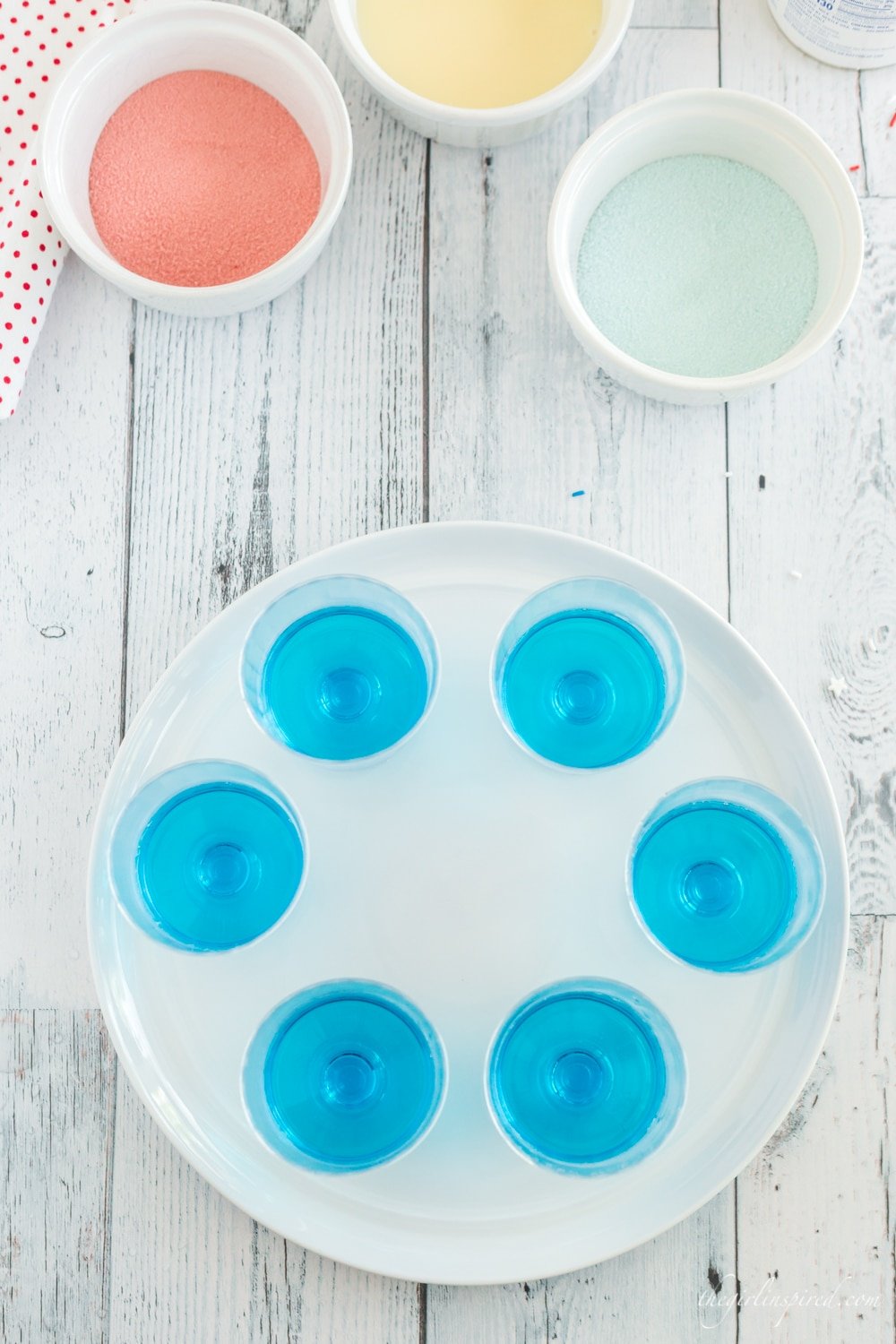 overhead photo of white plate with cups of blue jello, white bowls with red jello powder, blue jello powder, and sweetened condensed milk