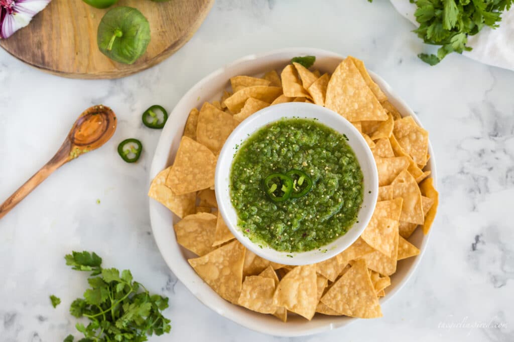 salsa verde in white chip'n'dip serving bowl with tortilla chips, wooden spoon, cilantro and jalapeño slices and wooden cutting board