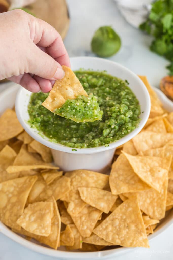 chip scooping salsa from white chip'n'dip bowl, tortilla chips, and a tomatillo and cilantro in background