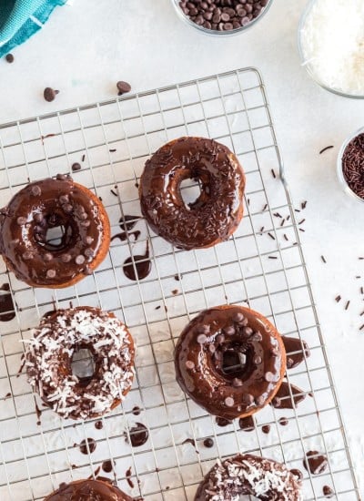 chocolate baked donuts on cooling rack with chocolate frosting dripping over the tops and coconut, chocolate chips, or sprinkles on top, aqua linen in background