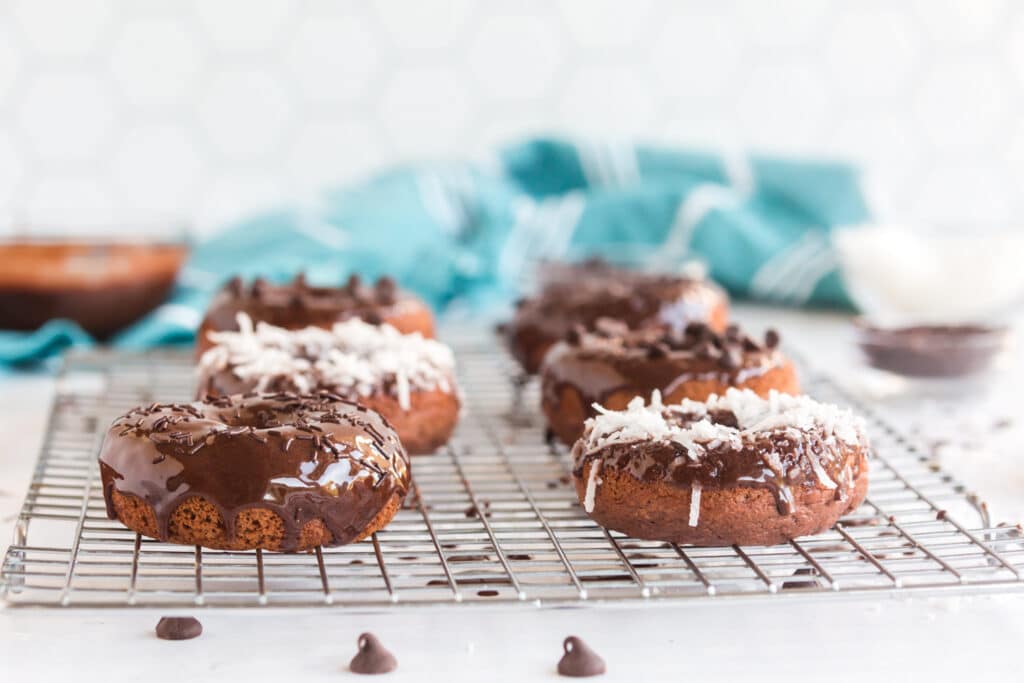 chocolate baked donuts on cooling rack with chocolate frosting dripping over the tops and coconut, chocolate chips, or sprinkles on top, aqua linen in background