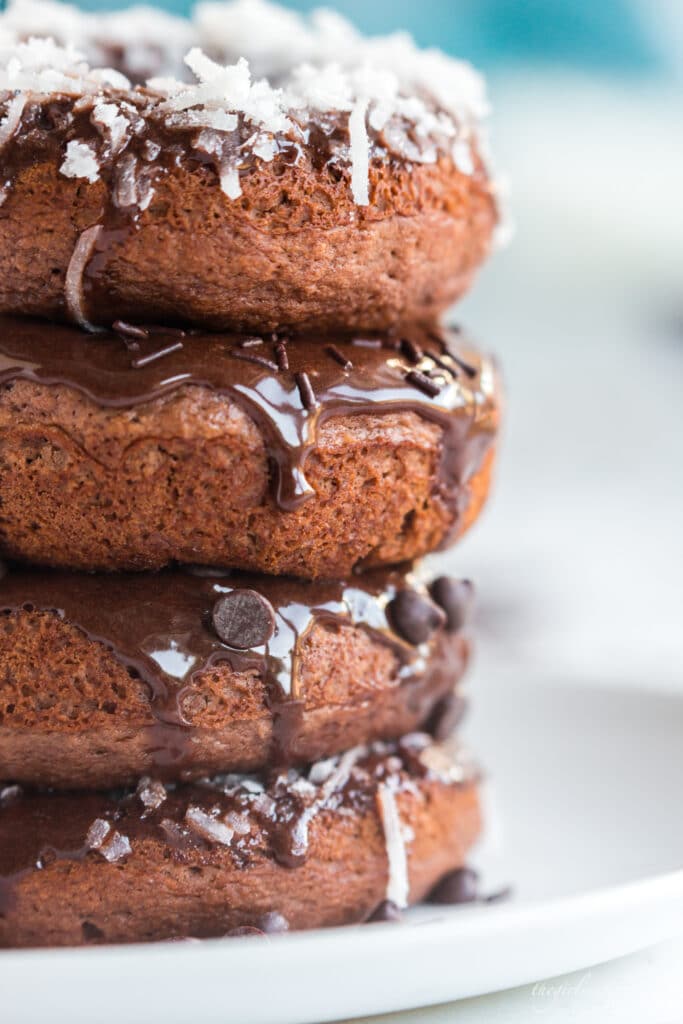 stack of chocolate baked donuts with chocolate frosting and toppings: coconut, sprinkles, and mini chocolate chips, on white plate