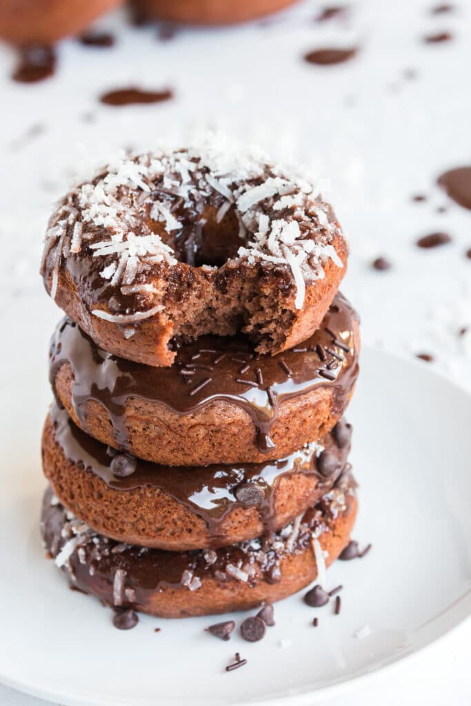 stack of chocolate baked donuts with chocolate frosting and toppings: coconut, sprinkles, and mini chocolate chips, on white cake platter