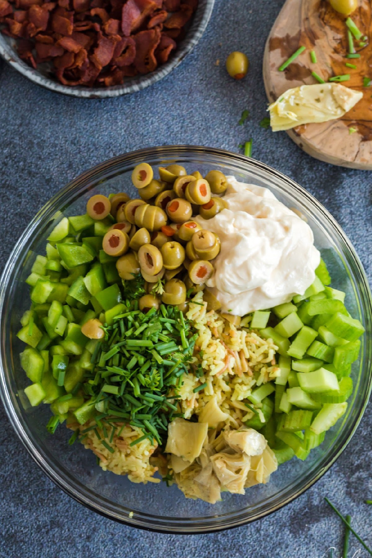 Glass bowl with piles of artichoke rice salad mixins mounded on top: mayo, celery, olives, bell pepper, artichoke hearts, and chives.