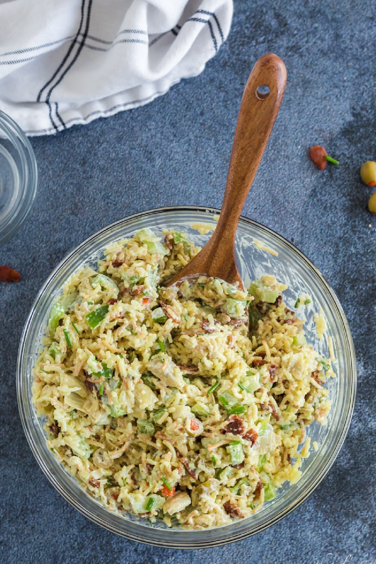 Glass bowl full of mixed up artichoke rice salad, wooden spoon, blue and white linen.