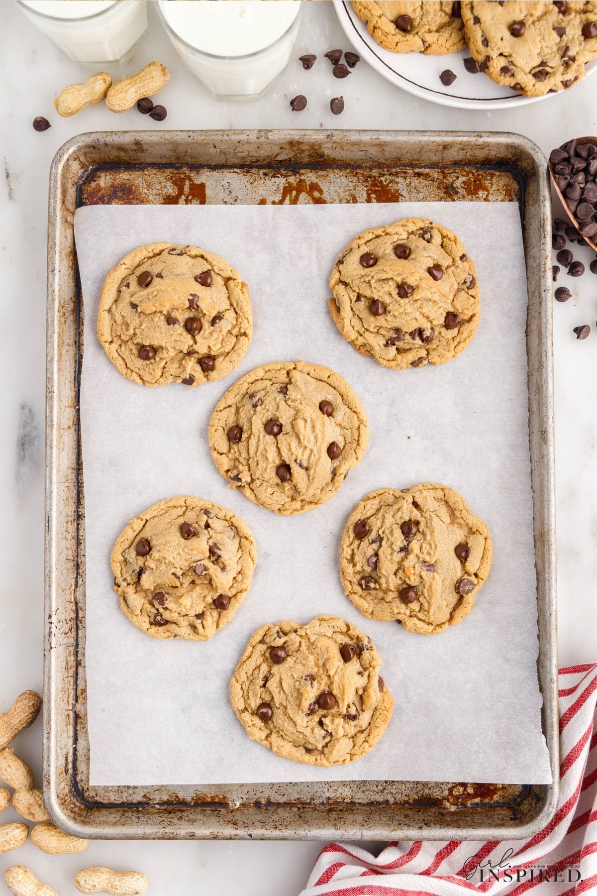 Baked peanut butter and chocolate chip cookies on a parchment-lined baking tray. Red and white striped cloth, glasses of milk, side plate of stacked cookies, and loose chocolate chips in the background.
