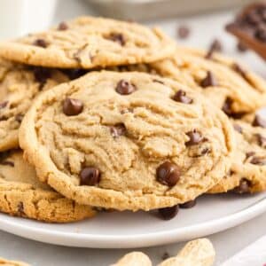 Large peanut butter chocolate chip cookies stacked on a plate with chocolate chips and whole peanuts scattered around.