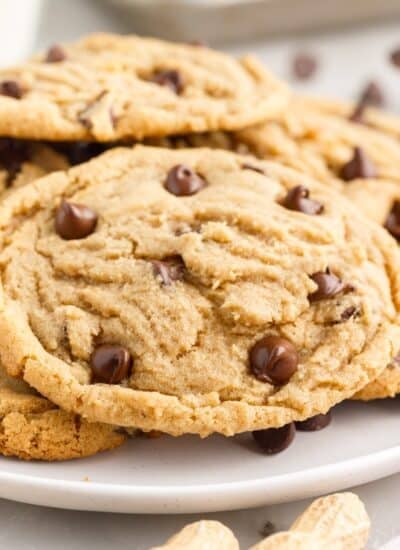 Large peanut butter chocolate chip cookies stacked on a plate with chocolate chips and whole peanuts scattered around.