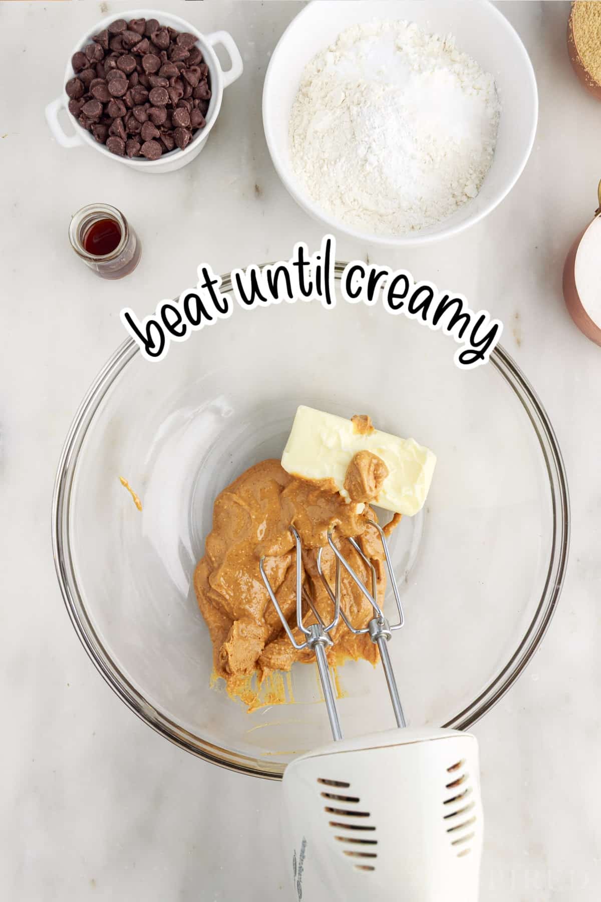 Mixing bowl containing softened butter and peanut butter with an electric hand mixer. Rest of the ingredients for the peanut butter chocolate chip cookies in the background.